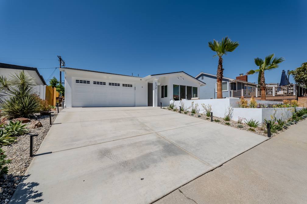 The finished front elevation on Avenida Cornelio: white single story house, wide concrete driveway, white stucco planters and palms.
