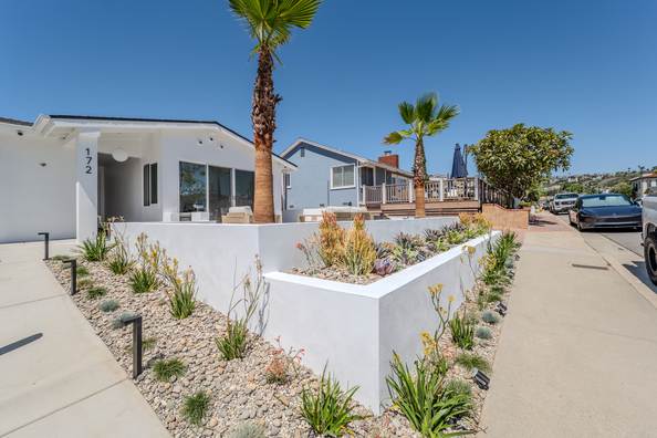 The house seen along the street, white planter wall and palms against a deep blue sky.