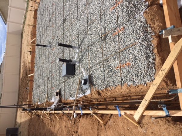 Gravel base with a rebar grid tied over it, edge forms set and plumbing stub-ups standing proud.