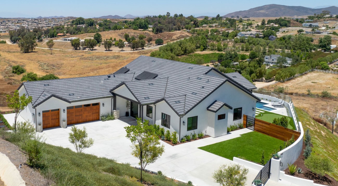 A finished custom home in Temecula wine country seen from the air: tile roof, three wood garage doors, poured driveway, lawn and pool, with the valley behind it.